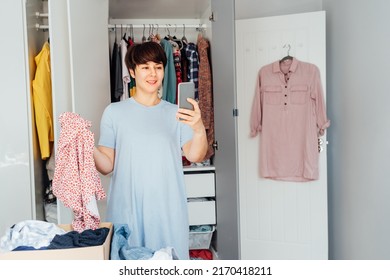 Smiling Woman In Bedroom Making A Video Call By Phone With A Stylist. Woman Selecting Clothes From Her Wardrobe. Decluttering, Sorting Seasonal Clothes And Cleaning Up. Fashion And Technology Concept.