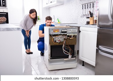 Smiling Woman Behind Technician Repairing Dishwasher In Kitchen
