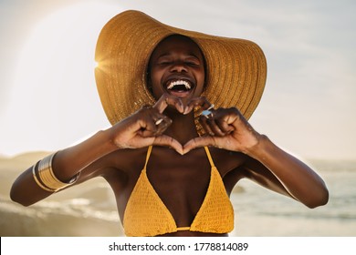 Smiling woman in bikini standing on the beach making a heart shape with her fingers. Female at the seashore wearing a sun hat making heart with hands.