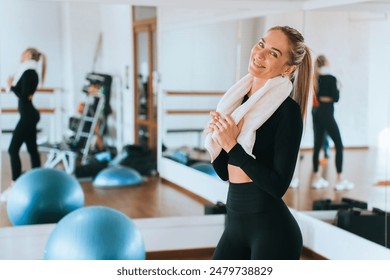 smiling woman in a black workout outfit with a white towel around her neck stands in a bright gym. She has long blonde hair tied back - Powered by Shutterstock