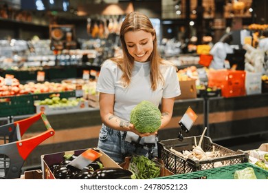 Smiling woman with blonde hair selects fresh savoy cabbage in a grocery store vegetable aisle, filling her cart with green, healthy options. Shopping concept - Powered by Shutterstock