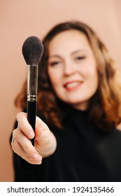 Smiling woman with blurred face showing her cosmetic brush, holding it in front of the camera. Selective focus on the object.