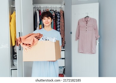 Smiling Woman with box of selected clothes from her wardrobe for donating to a Charity. Decluttering, Sorting clothes And Cleaning Up. Reuse, second-hand concept. Conscious consumer, sustainability