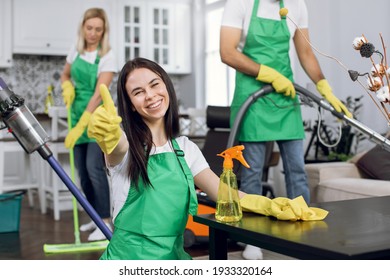 Smiling woman with brown hair showing thumb up on camera while her colleagues cleaning apartment. Professional housekeepers in uniform vacuuming, wiping and washing room.