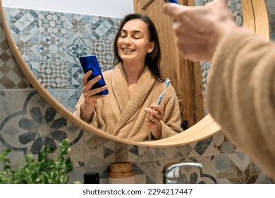 Smiling woman brushing teeth with toothbrush and using smartphone for chatting and blogging in front of mirror in bathroom in the morning. Oral hygiene, healthy teeth and care. - Powered by Shutterstock