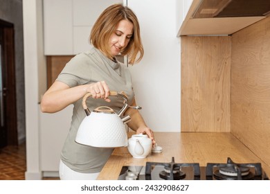 Smiling woman in a casual outfit pours boiling water from white kettle into teapot in modern kitchen setting. - Powered by Shutterstock