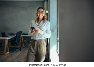 Smiling woman in casuals standing in office. Businesswoman with mobile phone in hand looking at camera. - Powered by Shutterstock