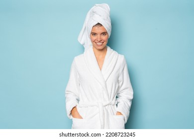 Smiling woman with clean body wearing white bathrobe and towel on head standing on blue studio background with hands in pocket at spa day on vacation or weekend. Hygiene, purity concept