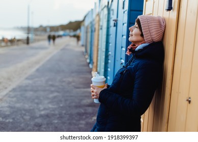 Smiling woman with closed eyes in warm clothes with a reusable cup with a hot drink near colorful beach booths and enjoying the moment. Simple pleasures and personal fulfillment. digital detox.