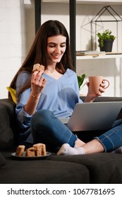 smiling woman with cup of coffee and piece of cake using laptop on sofa at home