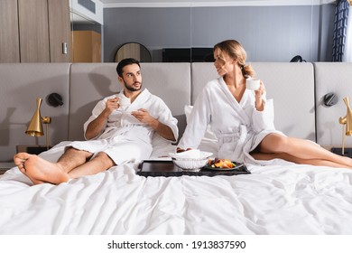 Smiling Woman With Cup Looking At Arabian Man Near Breakfast On Bed In Hotel