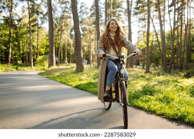 Smiling woman with curly hair in a coat rides a bicycle in a sunny park. Outdoor portrait. Beautiful woman enjoys nature. Lifestyle. Relax, nature concept.  - Powered by Shutterstock