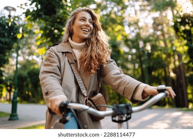 Smiling woman with curly hair in a coat rides a bicycle in a sunny park. Outdoor portrait. Beautiful woman enjoys nature. Lifestyle. Relax, nature concept.  - Shutterstock ID 2288114477