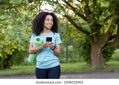 Smiling woman with curly hair holding smartphone and yoga mat in park. Engaged in fitness, enjoying outdoor environment, representing active and healthy lifestyle. Greenery and nature create peaceful - Powered by Shutterstock