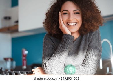 Smiling woman with curly hair leans on hand in a brightly lit kitchen. A blurred Christmas ornament in the foreground adds a festive touch. Her expression radiates warmth and joy - Powered by Shutterstock