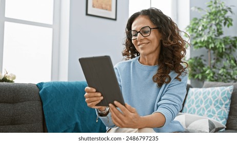 Smiling woman with curly hair wearing glasses and blue sweater using a tablet in a cozy living room - Powered by Shutterstock