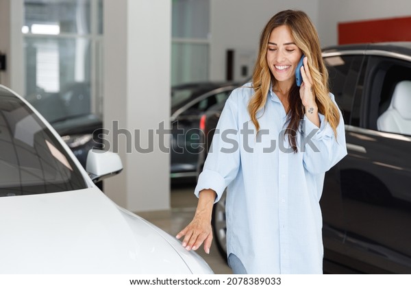Smiling woman customer female buyer client in\
blue shirt speak mobile cell phone chooses auto wants to buy new\
automobile in car showroom vehicle salon dealership store motor\
show indoor Sales\
concept