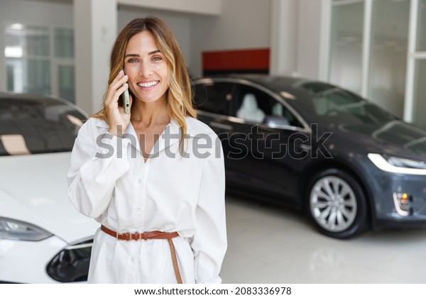 Smiling woman customer female buyer client in\
white shirt talk on mobile cell phone choose auto want buy new\
automobile in car showroom vehicle salon dealership store motor\
show indoor. Sales\
concept