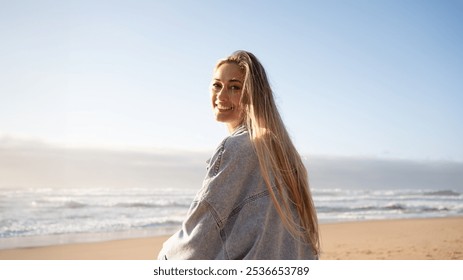 Smiling woman in denim shirt sitting on seashore against clear sky at beach during summer. Back view portrait of happy beautiful female with long blond hair is looking at camera.  - Powered by Shutterstock