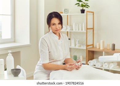 Smiling woman dermatologist sitting, holding moisturizing mask in jar and looking at camera over beauty salon interior at background. Professional cosmetologist at work