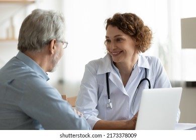 Smiling Woman Doctor Listening To Mature Patient Complaints At Meeting In Hospital, Friendly Therapist Physician Wearing White Uniform With Stethoscope Consulting Senior Man At Medical Appointment