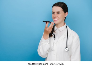 Smiling Woman Doctor Wearing White Medical Uniform And Stethoscope Holding Phone Speak Activate Virtual Digital Voice Assistant On Smartphone, Consult Patient Online, Posing Over Blue Background Wall