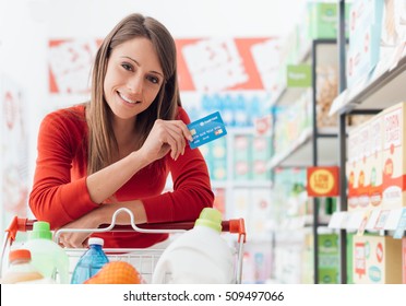 Smiling Woman Doing Grocery Shopping At The Supermarket She Is Leaning On A Full Cart And Holding A Credit Card