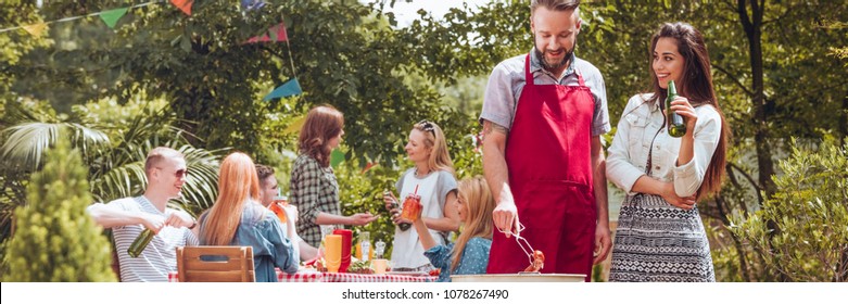 Smiling Woman Drinking Beer And Her Friend Grilling Food During A Meeting Outside