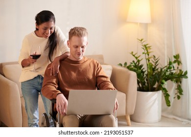 Smiling Woman Drinking Wine And Looking At Boyfriend Sitting On Wheelchair Working On Laptop