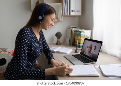 Smiling woman in earphones sit at desk at home look at laptop screen study distant with teacher, happy young female student make notes watch webinar or training course on computer, education concept