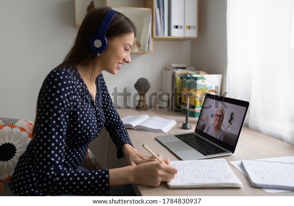 Smiling woman in earphones sit at desk at home\
look at laptop screen study distant with teacher, happy young\
female student make notes watch webinar or training course on\
computer, education\
concept