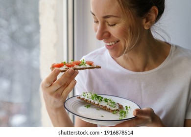 Smiling woman eating rye crisp bread with creamy vegetarian cheese tofu, cherry tomato and rucola micro greens, sitting at home and looking at window. Healthy food, gluten free, diet concept.  - Powered by Shutterstock