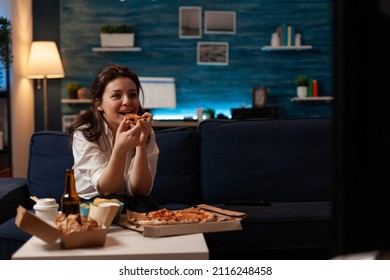 Smiling Woman Eating A Slice Of Hot Pizza Delivery Sitting On Couch Looking At Family Show On Television In Living Room. Happy Person Enjoying Takeaway Tv Dinner At Table With Takeout Fast Food.