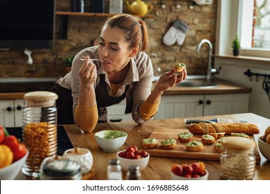 Smiling Woman Enjoying In Taste Of Healthy Food While Making Avocado Bruschetta In The Kitchen. 