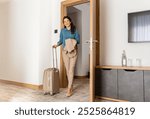 A smiling woman enters a hotel room, pulling her suitcase behind her and holding a jacket. She looks ready to settle in, with a modern, minimalist hotel setting in the background.