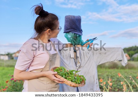 Smiling woman farmer with basket with spicy herbs hugging scarecrow in vegetable garden, nature landscape blue sky background