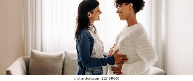 Smiling woman feeling a pregnant woman's belly. Happy young woman feeling the movement of a pregnant woman's baby. Cheerful young woman spending time with her surrogate at home.