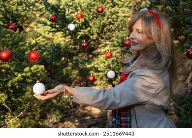 A smiling woman in a festive outfit adorns a pine tree with red and white Christmas ornaments - Powered by Shutterstock