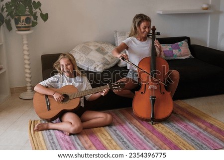 Smiling woman with fiddlestick sitting on sofa and playing cello while happy girl sitting on floor and playing guitar in living room at home
