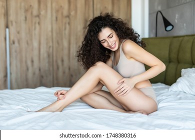 Smiling Woman With Fit Body And Beautiful Legs On White Bed. Happy Girl In White Underwear With Soft Depilated Body Skin, Natural Hair And Beauty Face In Light Interior. Women Skin Care. High Quality
