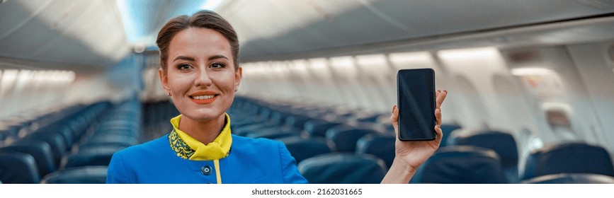 Smiling Woman Flight Attendant Holding Mobile Phone And Pointing At Passenger Seat While Standing In Aircraft Salon