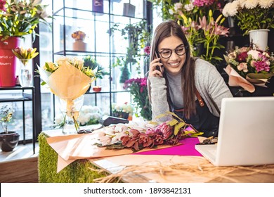 Smiling Woman Florist Small Business Flower Shop Owner. She is using her telephone and laptop to take orders for her store. Female gardener noting client order during mobile phone conversation