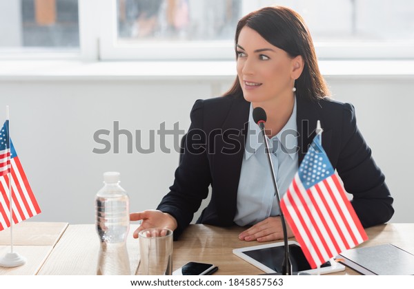 Smiling woman in\
formal wear looking away, while speaking in microphone, sitting\
near american flags in\
boardroom