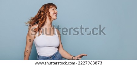 Smiling woman with ginger hair dancing and having fun against a blue background. Woman with arm tattoos celebrating while standing in a studio. Happy young woman wearing a tank top and jeans.