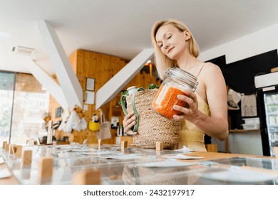 Smiling woman with glass jar of red lentils in zero waste shop. Female customer with basket of organic products in modern plastic free store. Concept of retail of eco food and sustainable shopping. - Powered by Shutterstock