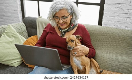 A smiling woman with grey hair cuddles her dog while using a laptop in a cozy living room interior. - Powered by Shutterstock