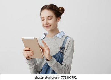 Smiling Woman With Hair Tied In Double Buns Looking At Screen Of Modern Device. Pleased Female Cutie Typing Message To Her Boyfriend Using IPad. Relationships Concept