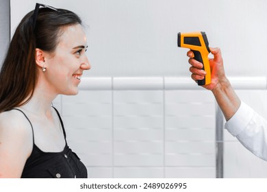 A smiling woman is having her temperature checked using an infrared thermometer held by a healthcare professional. The thermometer's laser is visible on her forehead. - Powered by Shutterstock