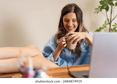 A smiling woman with a headset relaxing at her home office, feet up on the desk, and enjoying a coffee break. Casual and comfortable remote work setup - relaxed atmosphere, cozy home office. - Powered by Shutterstock