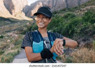 Smiling Woman Hiker In Cap Looking Away. Female Trail Runner Checking Smart Watch While Standing In Valley.